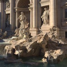 a fountain in front of a large building with statues on it's sides and water running down the side