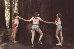 three girls are standing in front of a large tree holding hands with each other while wearing shorts