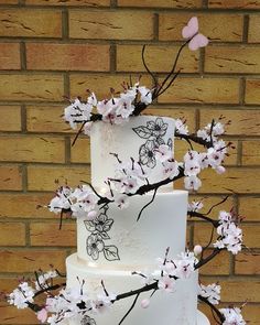 a three tiered white cake with cherry blossoms on the top and bottom, sitting against a brick wall