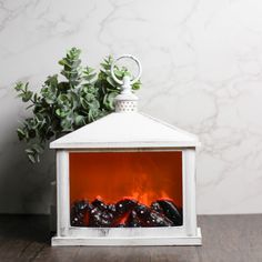 a white fireplace with an orange fire in it and some green plants on the side