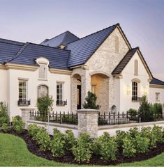 a large white house with a black roof and some plants in the front yard at dusk