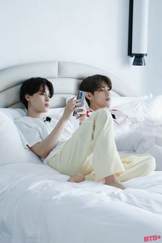 two people sitting on a bed with white sheets and one person holding a cell phone