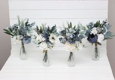 three vases with flowers and greenery are lined up against a white plank wall