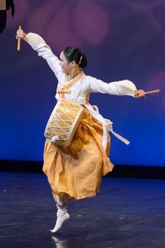 a woman in an orange dress is dancing on stage with a drum and stick,