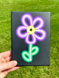 a hand holding up a purple and green flower on a black square with grass in the background