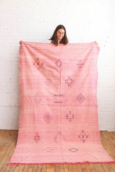 a woman holding up a pink blanket on top of a wooden floor in front of a white brick wall