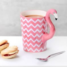 a pink flamingo mug next to two cookies and a spoon on a white table