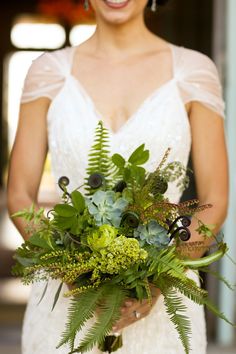 a woman in a wedding dress holding a bouquet of flowers and greenery on her arm