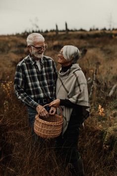 an older man and woman standing in tall grass with a basket on their shoulder, looking at each other