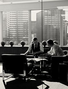 two men are sitting at a table in front of large windows with skyscrapers behind them