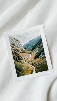 a card with a painting of a mountain road in the middle of it on a white sheet