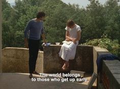 two people are standing on a ledge with trees in the background and one is holding a book