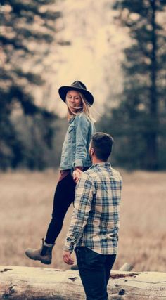 a man standing next to a woman on top of a log in a field with trees