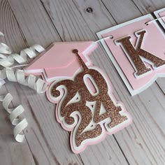 a pink and gold graduation cake topper with the number one on it, next to some white ribbons