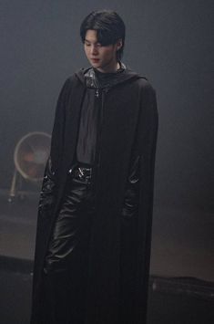 a male in a black coat and leather pants is standing on a stage with his hands behind his back