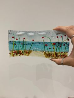 a person holding up a piece of paper with flowers and grass on it in front of the ocean