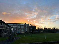 the sun is setting in front of a building with a soccer field and grass area