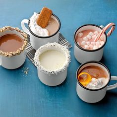 four mugs filled with hot chocolate, marshmallows and orange slices on a blue surface