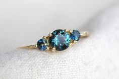 Trilogy ring featuring a bi-colour round brilliant cut sapphire with sapphire side stones on a fine tapered band with a polished finish. Teal Sapphire Ring, Teal Sapphire, Blue Gems