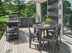 a table and chairs on a deck with an outdoor grill area in the back ground