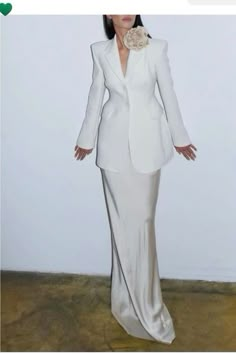 a woman wearing a white suit and flower brooch standing in front of a wall