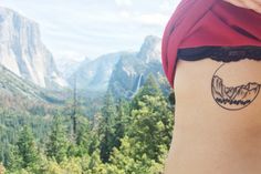 a woman with a tattoo on her stomach in front of some mountains and pine trees