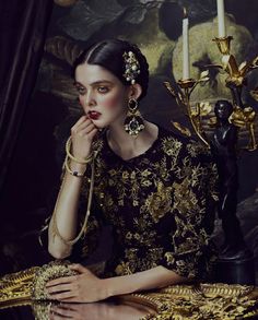 a painting of a woman sitting at a table with a candle in her hand and wearing gold jewelry