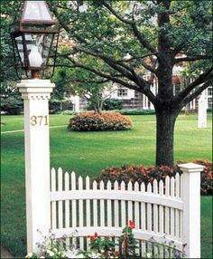 a white picket fence in front of a tree with flowers around it and a lamp post