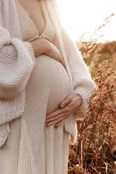 a pregnant woman standing in tall grass with her hands on her belly and wearing a white sweater