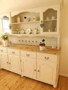 a white china cabinet with flowers and plates on the top, next to a wooden floor