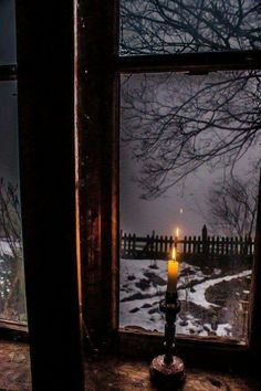 a candle is lit in front of an open window with snow on the ground and trees outside