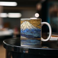 a coffee mug sitting on top of a table next to a cup with a mountain scene painted on it