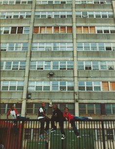 three boys are jumping on a fence in front of a tall building with windows and balconies