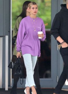 a woman in a purple sweater and white pants is walking down the street while holding a coffee