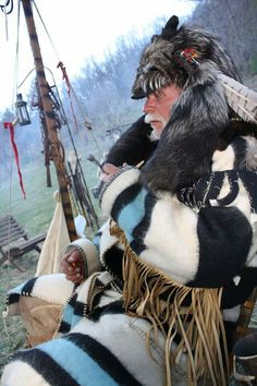 an old man sitting in a chair with a hat on his head and feathers around his neck