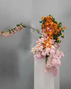 a white vase filled with lots of colorful flowers next to a gray wall and a pink ribbon