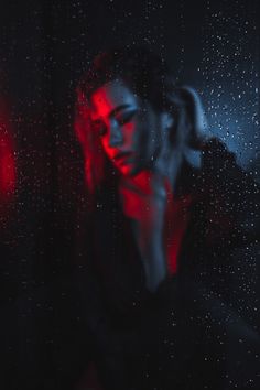 a woman standing in front of a window with rain drops on the glass and red light behind her