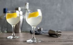 two wine glasses filled with white liquid and lemon wedges on a table next to bottles