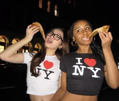 two young women eating pizza at night in front of a building with i love ny t - shirts on