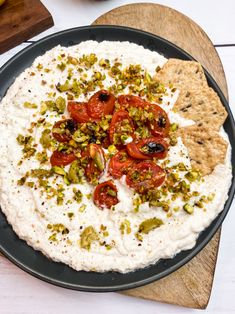 a black plate topped with hummus, tomatoes and pistachios next to crackers