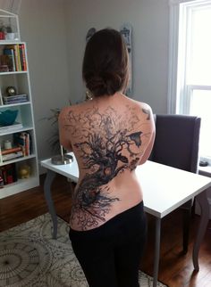 a woman with tattoos on her back standing in front of a table and bookshelf