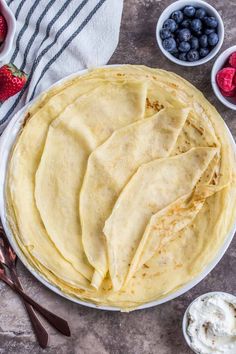 a plate filled with crepes next to bowls of strawberries and blueberries