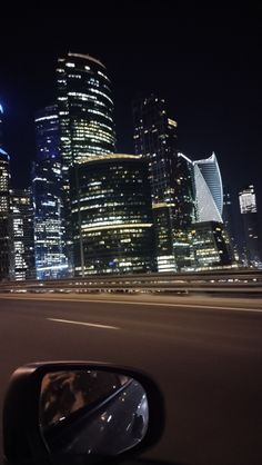 the city skyline is lit up at night with skyscrapers in the foreground and cars driving on the road