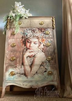 an ornate dresser with flowers on top and a painting on the front, sitting next to a window