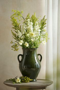 a green vase filled with white flowers sitting on top of a table next to a window