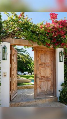 an open wooden door surrounded by greenery and pink flowers on the side of a building