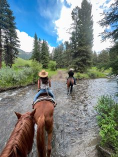 two people riding horses through a stream in the woods on a sunny day with blue sky and clouds