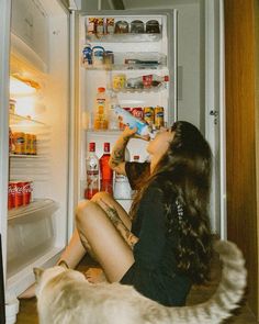 a woman is sitting in front of an open refrigerator drinking from a bottle while a dog looks on