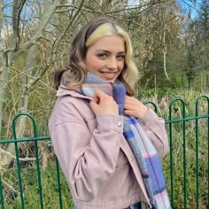 a woman wearing a pink jacket and blue plaid scarf standing in front of a fence