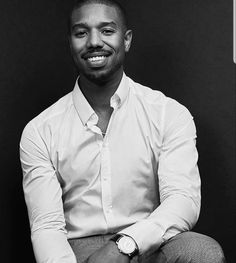 a black and white photo of a man in a dress shirt sitting on a stool